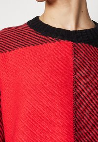 Gros plan d'une personne portant un pull en tricot texturé rouge et noir avec un col côtelé noir, montrant le cou et la partie supérieure de la poitrine.