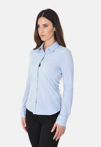 Camicia azzurra a maniche lunghe con colletto a punta, bottoni frontali e design aderente. Il tessuto appare liscio con una leggera lucentezza. Abbinata a pantaloni neri.