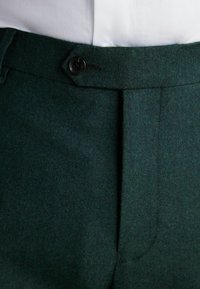 Tummanvihreät villahousut, joissa on teksturoitu pinta, etutaskut pleatilla ja musta nappi vyötäröllä.
