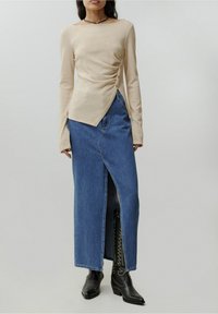 Beige Langarmoberteil mit seitlicher Raffung und Knopfdetails; kombiniert mit einem blauen Jeansmaxirock mit seitlicher Schlitze und schwarzen Ankle Boots.