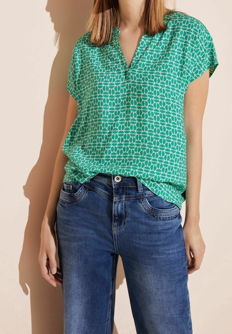Zielona bluzka z krótkim rękawem w geometryczny wzór, z dekoltem w kształcie V, w zestawie z niebieskimi dżinsami.