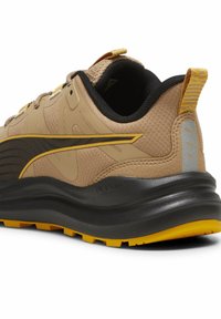 Tan athletic shoe with textured fabric, black accents, and yellow highlights. Features a cushioned sole and visible brand logo on the side.