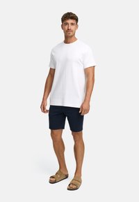 Homme debout portant un t-shirt blanc uni, un short bleu marine et des sandales beiges sur un fond gris clair uni.