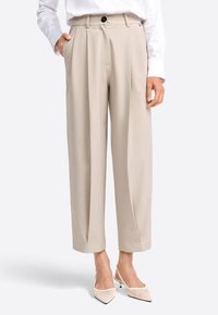 Pantaloni beige a gamba ampia con pieghe frontali, passanti per cintura e tasche laterali, abbinati a décolleté beige con cinturino sullo sfondo neutro.