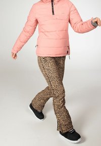 Personne portant une veste matelassée rose, un pantalon à imprimé léopard et des baskets noires avec des semelles blanches, marchant sur un fond clair uni.