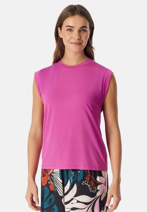 Vrouw draagt een mouwloze felroze top en een broek met een bloemenpatroon, staand tegen een effen witte achtergrond.