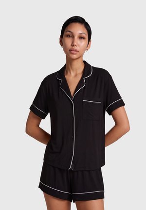 Femeie cu păr scurt și închis la culoare, purtând un top negru de pijamale, cu mâneci scurte și nasturi și pantaloni scurți asortați cu margine albă, stând cu mâinile la spate.