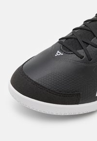 Černá sportovní bota s hladkým syntetickým svrškem, texturovanými akcenty a bílou gumovou podrážkou s tečkovaným vzorem pro lepší přilnavost.