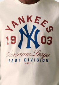 Camiseta crema con texto en rojo y azul en bold. El diseño incluye "Yankees", "1903", "Liga Americana" y "División Este" con un logo.