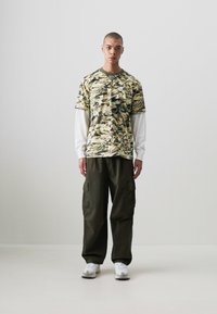 Camo-patterned short-sleeve shirt over a white long-sleeve shirt, paired with dark green cargo pants and white sneakers.