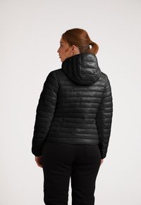 Giacca a vento nera con cappuccio, caratterizzata da un design trapuntato e una texture a costine. La vista posteriore mostra cuciture orizzontali e una vestibilità aderente.