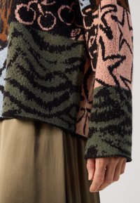 Gros plan sur la main et le bras d’une personne portant un pull texturé avec des motifs abstraits verts, noirs, roses et marron sur une jupe vert olive.