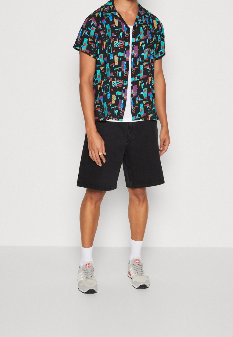 Kortebloes met een meerkleurig abstract patroon op een zwarte achtergrond, gecombineerd met zwarte shorts tot de knie en grijze sneakers.