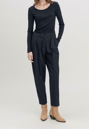 Vrouw draagt een nauwsluitend zwart shirt met lange mouwen, hoge taille plissé zwarte broek en beige enkellaarzen tegen een lichte achtergrond.