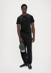 T-shirt preta de mangas curtas com gola em borgonha, combinada com calças pretas. Boné preto na mão com o logótipo "Couture" e ténis pretos.