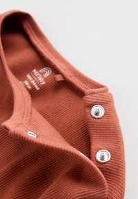 Gros plan d'un tissu côtelé de couleur rouille avec deux pressions argentées sur l'encolure d'un vêtement.