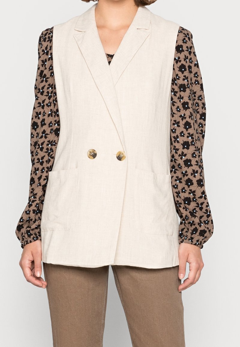 Gilet en lin beige à double boutonnage, deux poches avant. Associé à un chemisier marron à imprimé floral noir et manches bouffantes.