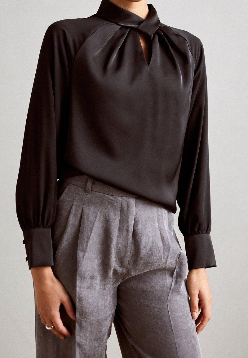 Femme portant un chemisier noir à manches longues avec un col « keyhole » et un pantalon plissé gris clair taille haute.