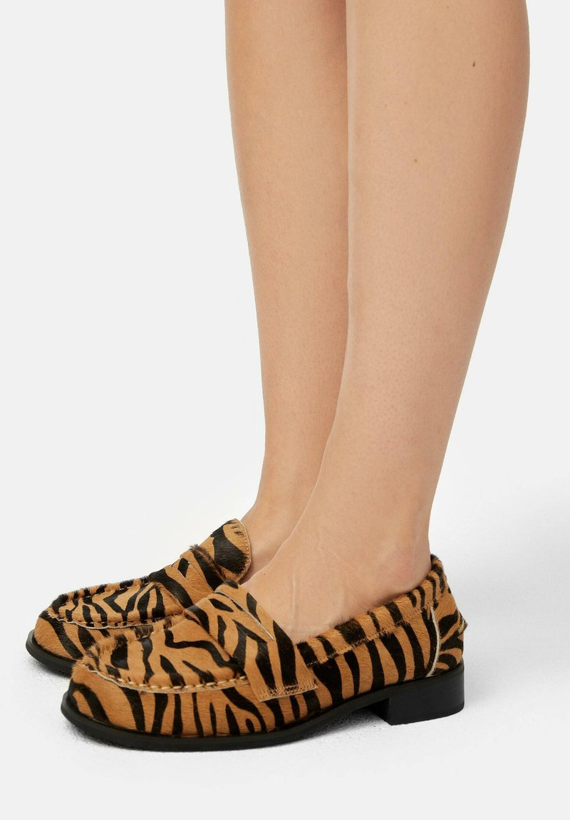 Loafers w paski w kolorze brązowym i czarnym, wykonane z teksturowanego materiału. Mają niski obcas i miękką cholewkę z zaokrąglonym noskiem.