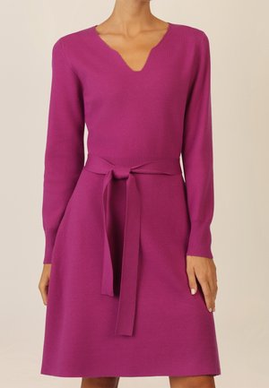 Kobieta ubrana w długą sukienkę z długim rękawem w kolorze magenta, z dekoltem w kształcie litery V i wiązanym paskiem w talii, na jednolitym jasnym tle.