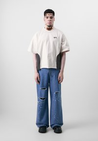 Beige übergroßes T-Shirt mit schwarzen Akzenten, kombiniert mit ausgewaschenen blauen Schlagjeans mit Farbspritzern. Schwarze klobige Schuhe.