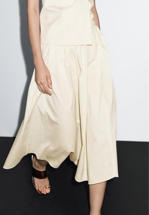 Kvinna som går i lös, krämfärgad plisserad kjol och ärmlös topp, bär mörkbruna platta sandaler med tåring på svart golv.