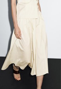 Woman walking in loose, cream-colored pleated skirt and sleeveless top, wearing dark brown flat sandals with a toe ring on black floor.