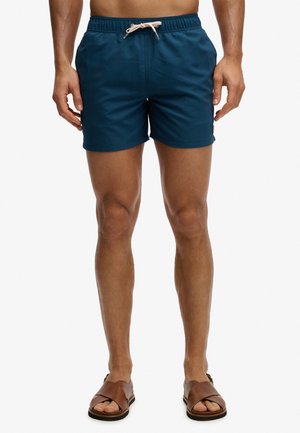 Mężczyzna w granatowych kąpielówkach ze białym sznurkiem i brązowych skórzanych sandałach stoi na białym tle.