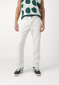 White denim jeans with a straight-leg cut, five pockets, and stitching details, paired with black and green sneakers.