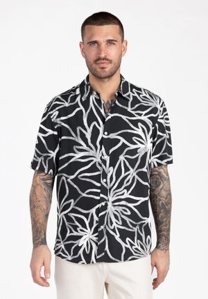 Uomo con capelli corti e tatuaggi che indossa una maglietta nera a maniche corte con grandi motivi floreali bianchi, in piedi contro uno sfondo bianco.