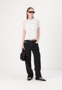 T-shirt bianca a maniche trasparenti, abbinata a jeans neri a vita alta. Gli accessori includono una piccola borsa nera e delle scarpe ballerine a punta.