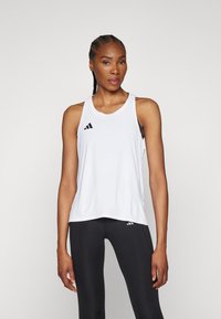 Bijela sportska majica bez rukava s izrezom u obliku uvale, s crnim Adidas trostrukim logotipom na lijevoj strani prsiju, u kombinaciji s crnim tajicama.