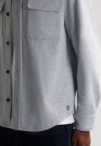 Szara flanelowa koszula z zapięciem na guziki z przodu, dwoma kieszeniami na piersi i zaokrąglonym dołem. Posiada ciemne guziki oraz małe logo w pobliżu dołu.