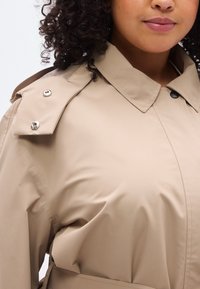 Μπεζ αδιάβροχο παλτό με ψηλή γιακά, κουμπιά στους ώμους και ζώνη στη μέση. Λεία υφή με καθαρό, μοντέρνο σχεδιασμό.