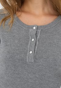 Gros plan sur une personne portant un t-shirt en maille grise avec un col rond et trois boutons blancs sur la patte avant.