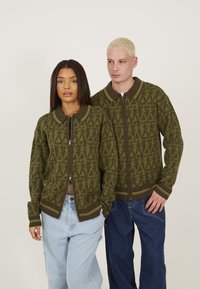 Olive green zip-up cardigan with patterned texture, brown collar, and accent stripes. Worn by two individuals with relaxed-fit jeans.