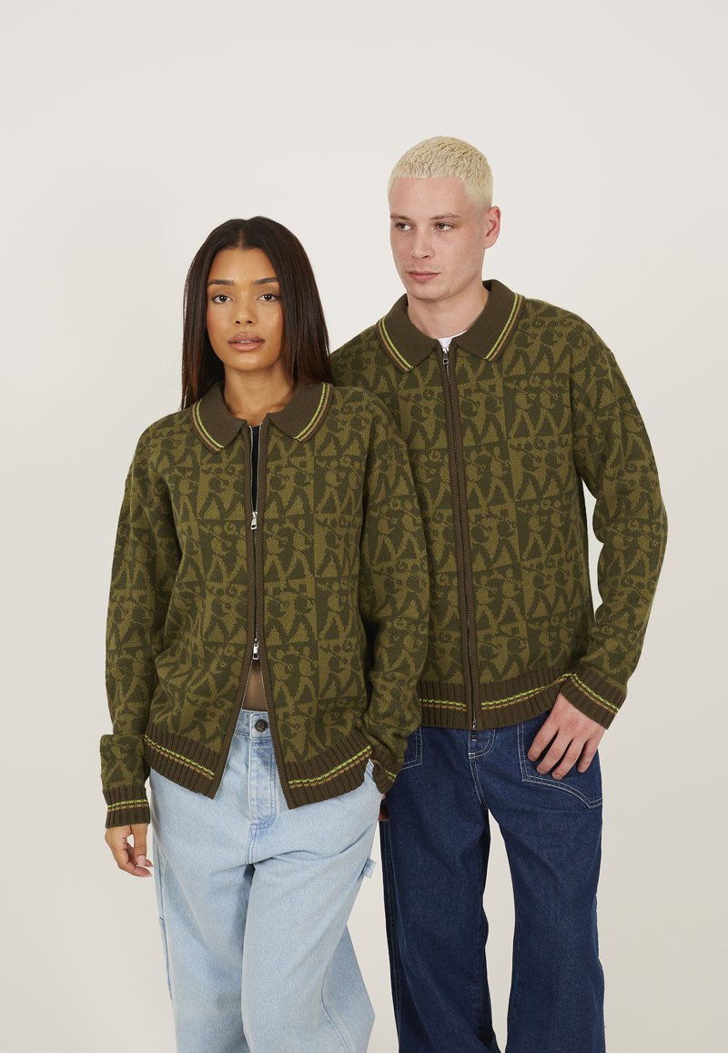 Olive green zip-up cardigan with patterned texture, brown collar, and accent stripes. Worn by two individuals with relaxed-fit jeans.