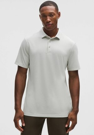 Junger Mann in einem hellgrauen, kurzärmligen Poloshirt und dunklen Hosen, der vor einem schlichten hellen Hintergrund steht.