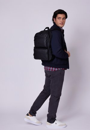 Mochila negra con múltiples bolsillos, textura suave y correas ajustables, llevada sobre una chaqueta azul marino y una camisa de cuadros con pantalones grises.