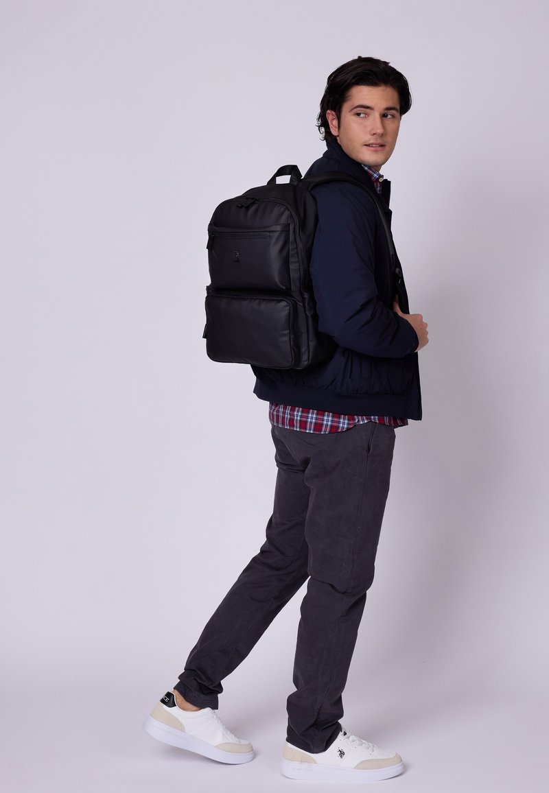 Zaino nero con molte tasche, texture liscia e spallacci regolabili, indossato sopra una giacca blu navy e una camicia a quadri con pantaloni grigi.
