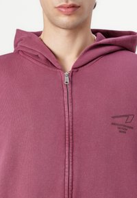 Hanorac cu fermoar de culoare vișinie, fabricat din bumbac moale. Prezintă o capotă și un mic logo pe partea stângă, având o textură netedă și o croială lejeră.