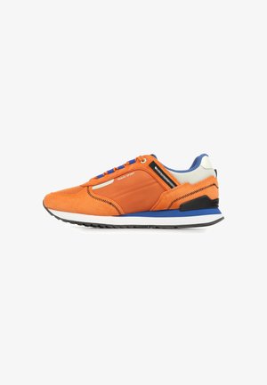 Oranje lage sneaker met blauwe en witte zool, blauwe veters, zwarte zigzagstiksels en de tekst "TRAVIS SPORT" aan de zijkant.