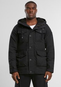 Sort parka-jakke med hætte, der har en Sherpa-fôr, flere forlommer, knaplukninger og justerbare manchetter.