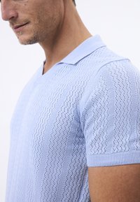 Světle modrá pletená polokošile s krátkými rukávy, zdobená strukturovaným vzorem vertikálních linií a průsvitnými detaily, žebrovaným límcem a manžetami.