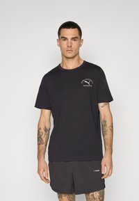 Sort bomulds T-shirt med rund hals og korte ærmer, prydet med et lille hvidt logo på brystet. Bæres med mørke shorts.
