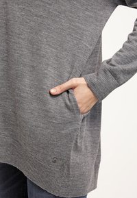 Personne portant un pull gris en tricot avec une main insérée dans la poche latérale, montrant les détails des poignets et de l'ourlet côtelés.