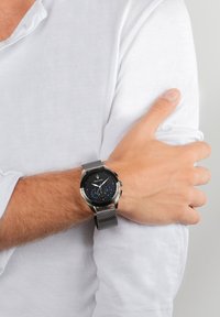 Svart och silverklocka med ett mesharmband, rund urtavla med blå accenter och silverdetaljer, bärs på ett handled med en vit skjorta.