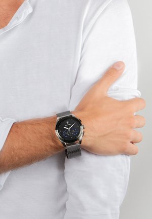 Zwarte en zilveren horloge met een mesh-band, ronde wijzerplaat met blauwe accenten en zilveren hardware, gedragen om een pols met een wit shirt.