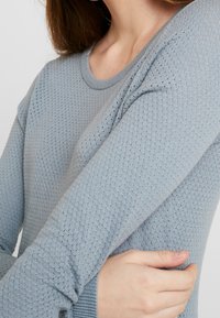 Hellblauer, strukturierter Langarmpullover mit Rundhalsausschnitt und engen Bündchen, der ein diamantförmiges Muster im gesamten Stoff aufweist.
