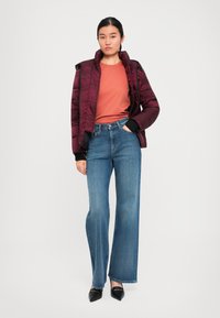 Young woman wearing a maroon puffer jacket, orange top, blue wide-leg jeans, and black pointed shoes standing with one hand in pocket.